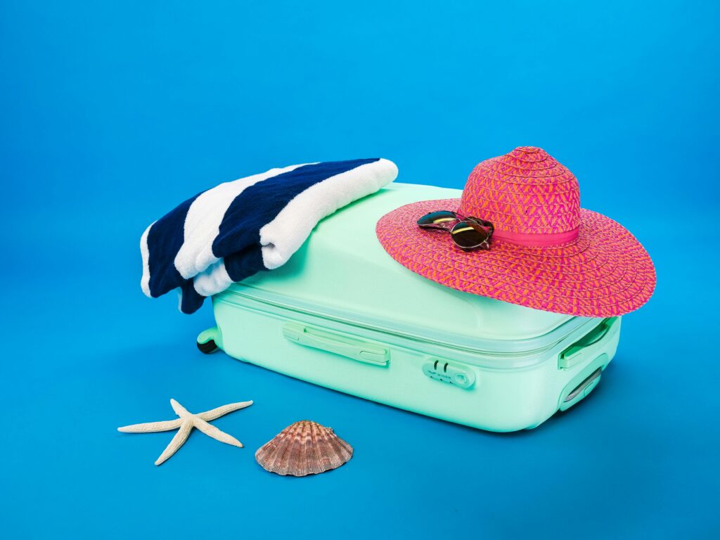 pexels-photo-8185803-8185803 Beach essentials including a suitcase, hat, towel, and seashells on a bright background.